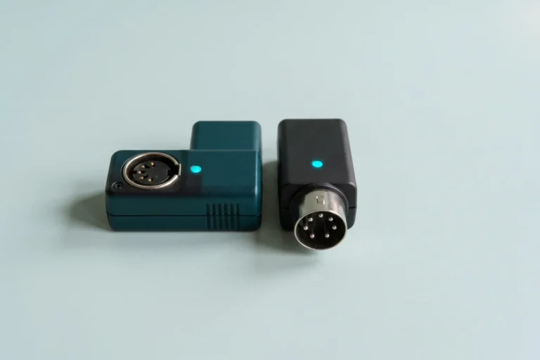 A teal and charcoal pair of compact Bluetooth MIDI adapters with five-pin DIN connectors and small cyan status lights sits on a pale mint-gray surface, illustrating the best Bluetooth MIDI adapters.