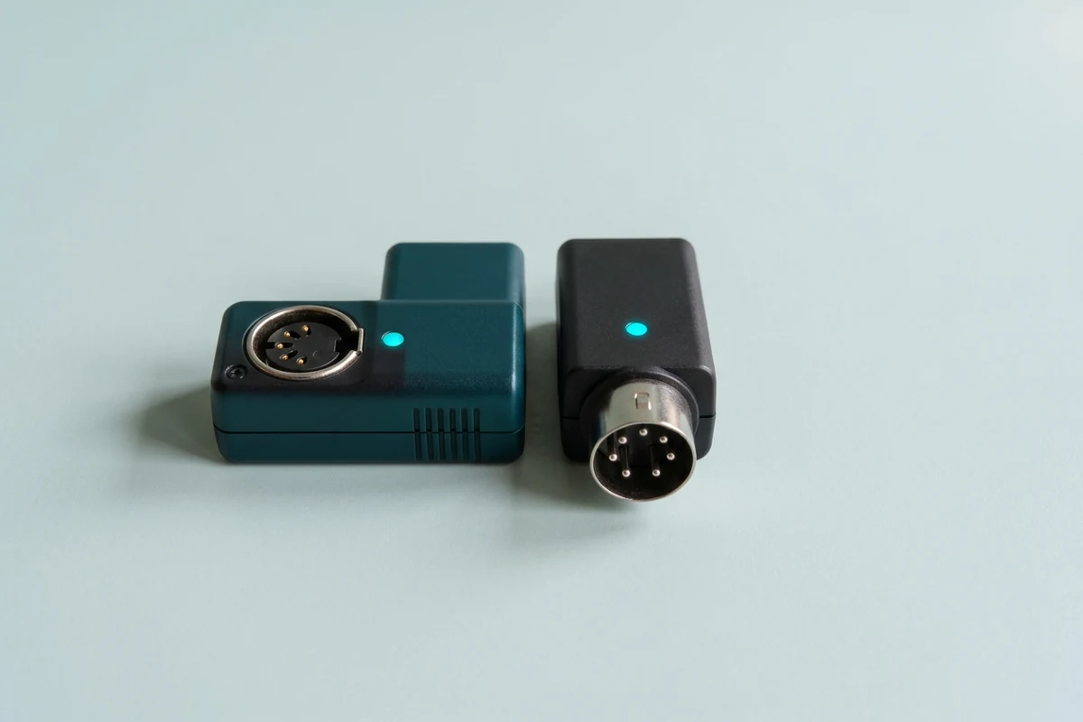 A teal and charcoal pair of compact Bluetooth MIDI adapters with five-pin DIN connectors and small cyan status lights sits on a pale mint-gray surface, illustrating the best Bluetooth MIDI adapters.